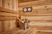 The Almost Heaven BlackWater 4 Person Cube Sauna features a cedar interior with a metal bucket and ladle on the bench. Two round gauges above the bucket display temperature and humidity, highlighting classic sauna comfort.