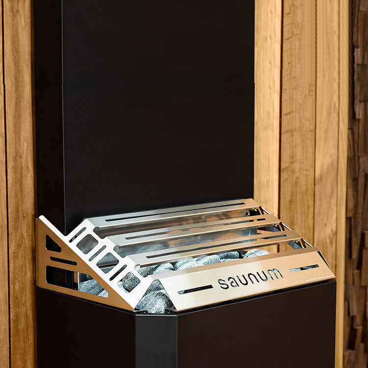 The Saunum AIR 7 WiFi Sauna Heater Package by Saunum, with a black body and stainless steel cover, features an innovative air blending system and sauna stones, and is installed against a wooden wall.