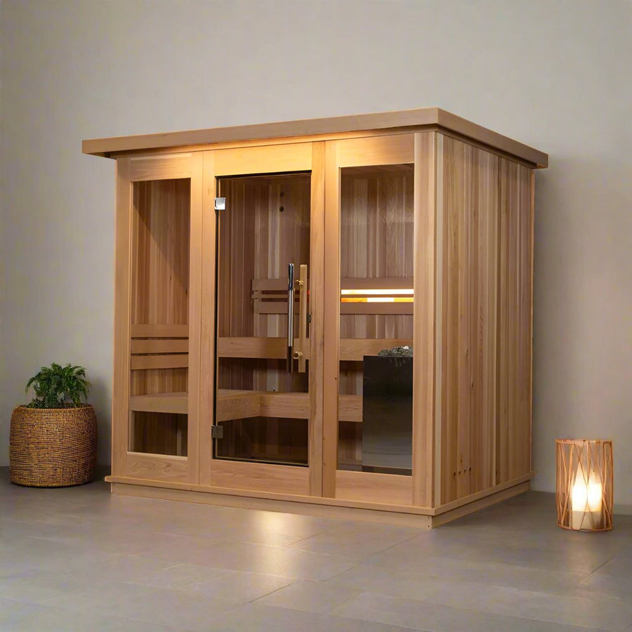 The Almost Heaven Denali 6 Person Indoor Sauna features modern cedar design with a glass door and panels, set in a minimalist room with light tile flooring, a plant in a woven basket on the left, and a softly lit wooden lantern on the right.