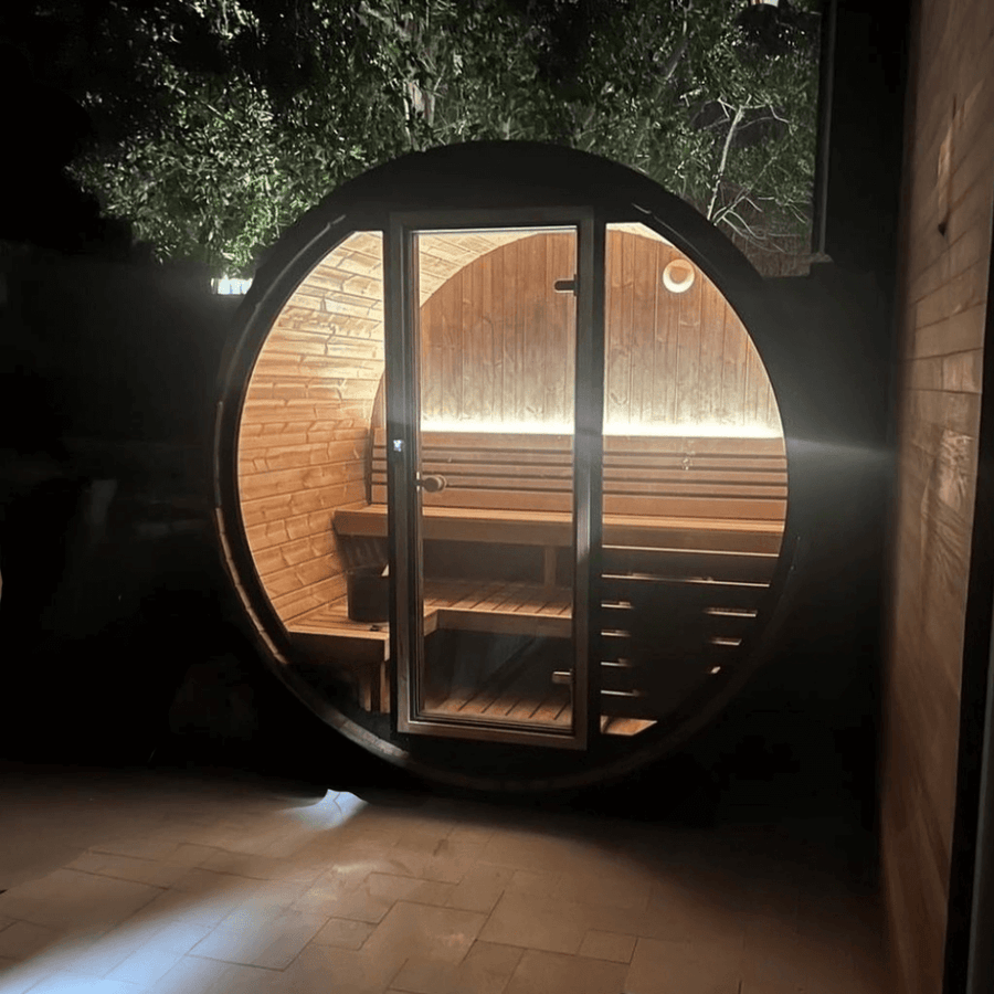 The SaunaLife Model EE6G 4 Person Barrel Sauna glows warmly at night, its glass door showing curved benches inside. Nestled outdoors beneath leafy trees, this inviting SaunaLife retreat is the perfect home sauna experience.