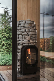 The HUUM Hive Flow LS Wood Burning Sauna Stove, a modern and eco-friendly heater with glass door and stone-filled metal frame, stands on a wooden floor inside a glass-walled room, with greenery and stacked firewood outside.