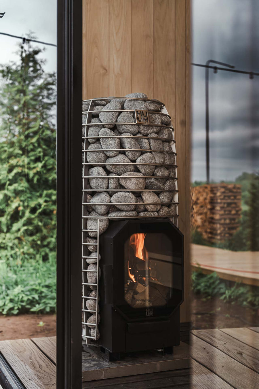 The HUUM Hive Flow LS Wood Burning Sauna Stove, filled with grey stones, stands in a wooden room with a glass wall revealing greenery and firewood outside as a small fire burns inside the HUUM stove.