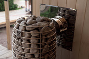 The HUUM Hive Flow LS Wood Burning Sauna Stove features a metal basket of smooth stones attached to a pipe and wall, set against wooden walls with a view outside—combining eco-friendly design and premium quality by HUUM.