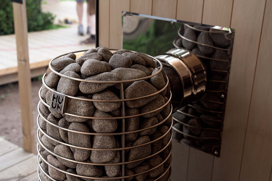 A metal basket of sauna stones is mounted on a HUUM Hive Flow LS Wood Burning Sauna Stove, attached to the wall inside a wooden sauna room. The heater features a shiny metal vent and "HUUM" branding on its side.