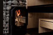 The HUUM Hive Flow LS Wood Burning Sauna Stove by HUUM features sauna stones atop a metal frame, a visible bright flame behind its glass door, and light wooden benches in the background, offering a modern and eco-friendly sauna experience.