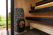 A modern indoor sauna with wooden benches features the HUUM Hive Flow LS Wood Burning Sauna Stove by HUUM, showcasing visible flames and stones. A ladle and bucket rest on the top bench. Natural light enters through an open glass door, revealing greenery outside.
