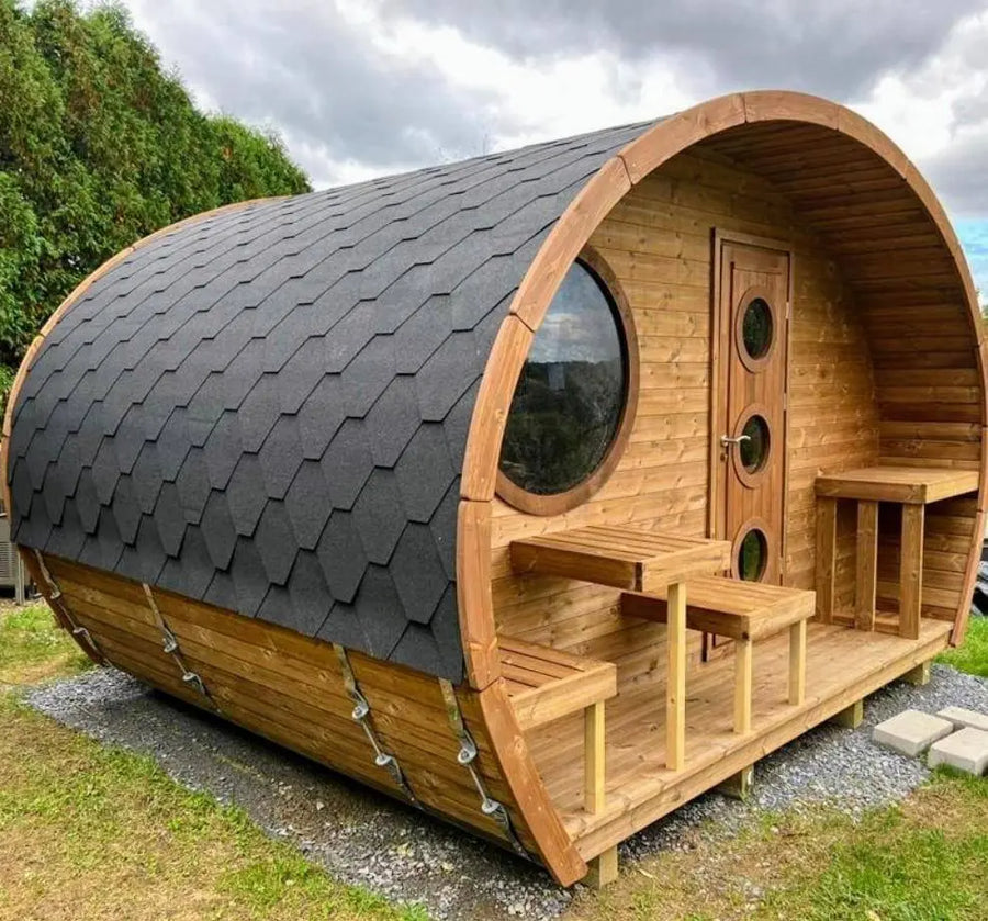 The SaunaLife Model G11 Outdoor 8 Person Sauna by SaunaLife features a barrel-shaped design, curved black shingled roof, round window, and a cozy porch with table and benches, set on gravel and grass under a cloudy sky.