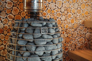 A close-up of the HUUM Hive Flow LS Wood Burning Sauna Stove, filled with smooth gray stones in a metal HUUM-branded cage, set against a decorative wall featuring a wood log cross-section pattern.