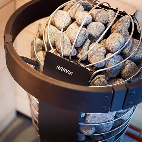 Close-up of a Harvia metal sauna heater filled with large round stones and surrounded by light wood walls. A Harvia Spirit Safety Rail provides added protection around the popular Harvia Spirit Series Heater.