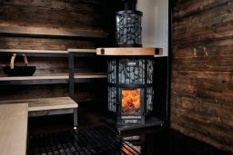 The Harvia Legend 240 GreenFlame Sauna Stove by Harvia is a modern wood-burning sauna stove with a glass door showing the fire, set against dark wooden walls and benches; a basket rests on a shelf in the background, offering an authentic sauna experience.