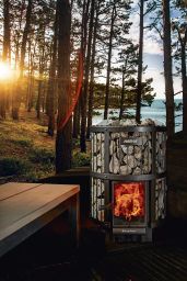 A Harvia Legend 240 GreenFlame Sauna Stove by Harvia, glowing with fire, stands on a deck in a forest clearing, sunlight filtering through tall trees and a lake visible in the background.