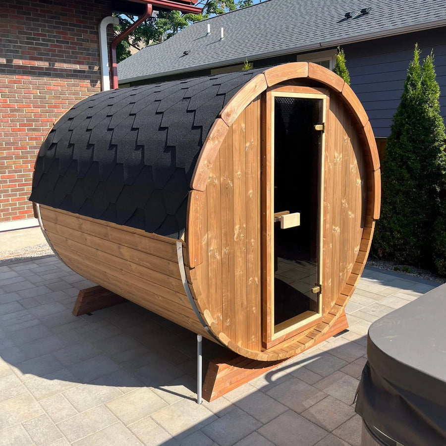 A SaunaLife Model E6W 3 Person Sauna Barrel-Window sits on a paved patio by a brick wall and green bushes, featuring a shingled black roof and a glass door with wooden handle.