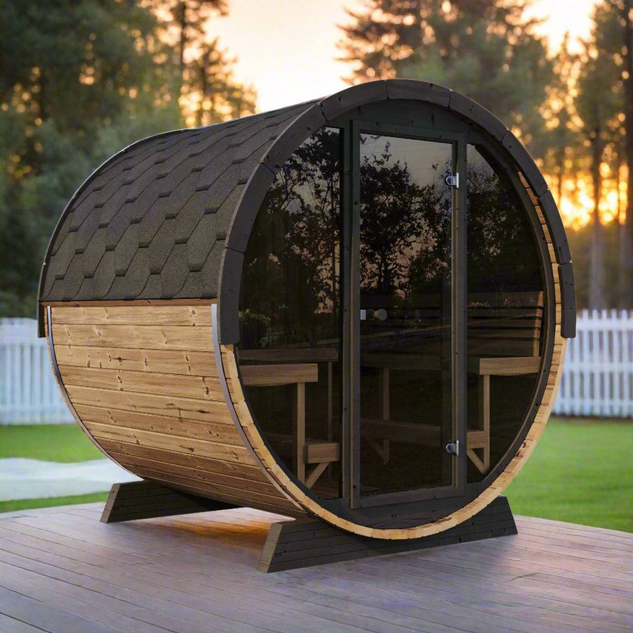 The SaunaLife Model EE8G 6 Person Barrel Sauna, featuring dark shingles and glass doors, sits on a wooden deck in a backyard surrounded by trees and a white picket fence at sunset.