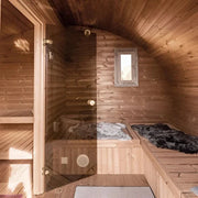 The SaunaLife Model G11 Outdoor 8 Person Sauna by SaunaLife features curved wooden walls and ceiling, a small window, wooden benches with soft fur throws, and a glass door for a cozy and inviting backyard retreat.