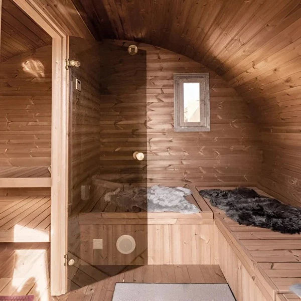 The SaunaLife Model G11 Outdoor 8 Person Sauna by SaunaLife features curved wooden walls and ceiling, a small window, wooden benches with soft fur throws, and a glass door for a cozy and inviting backyard retreat.
