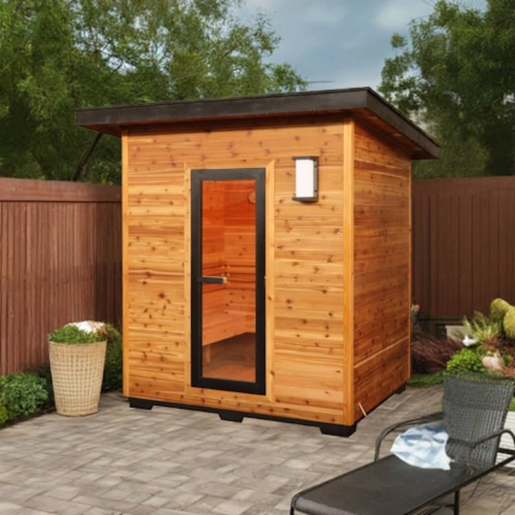 The SunRay Denali 4-Person Contemporary Outdoor Sauna by SunRay features a modern red cedar build, glass door, and sits on a patio surrounded by greenery and a lounge chair—ideal for a relaxing outdoor sauna experience.