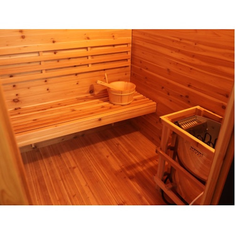 The SunRay Denali 4-Person Contemporary Outdoor Sauna features a spacious red cedar interior with a bench along one wall, a wooden bucket and ladle set on the bench, and a sauna heater positioned in the right corner.