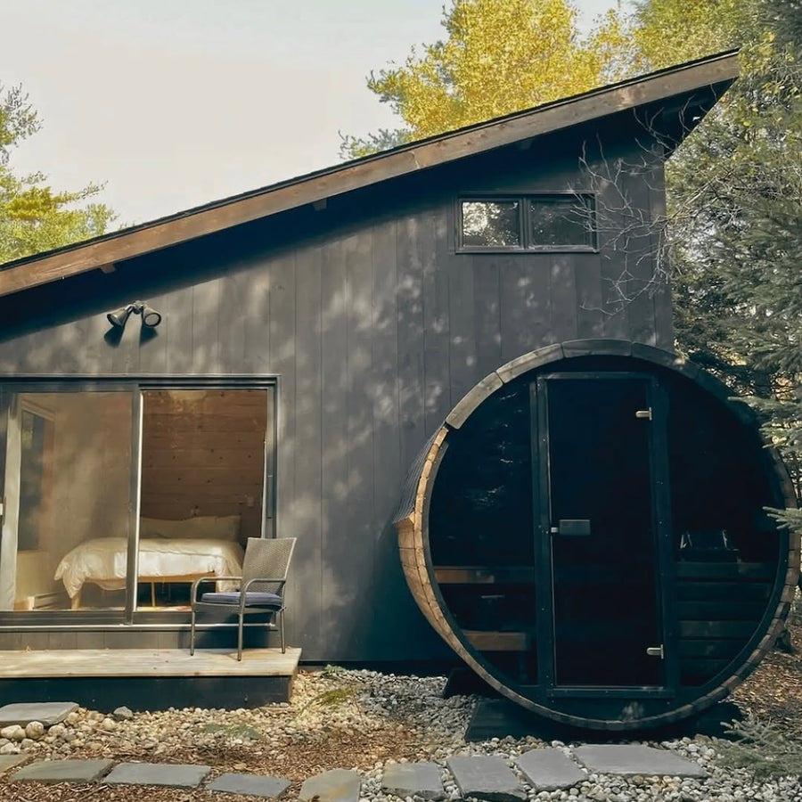 A modern black cabin with large windows and a small porch sits among trees. Next to it is the SaunaLife Model EE6G 4 Person Barrel Sauna by SaunaLife. Inside, a neatly made bed is visible. Stones line the path in front of the building.