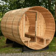 The Almost Heaven Charleston 4 Person Canopy Barrel Sauna by Almost Heaven features a cylindrical wooden design, glass door, benches, and a Harvia 6kW heater, set outdoors on grass and surrounded by trees.