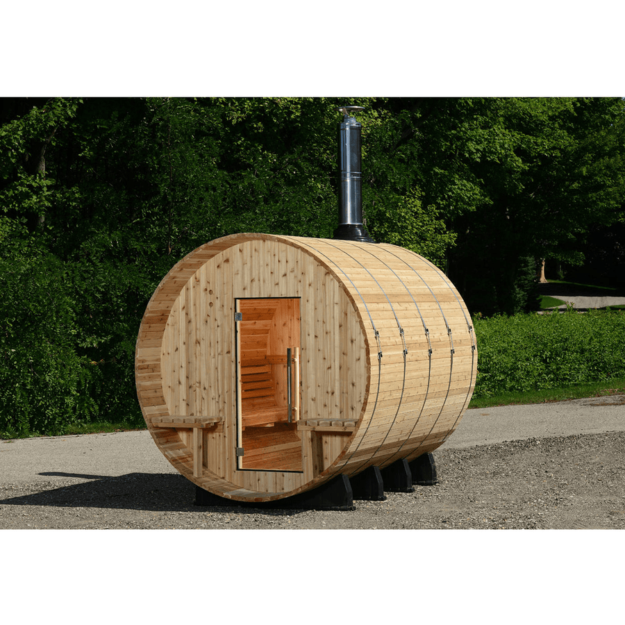An Almost Heaven Grandview 4-6 Person Canopy Barrel Sauna, distinguished by its round barrel shape, glass door, and chimney, is placed outdoors on gravel among green trees with built-in benches outside the entrance.