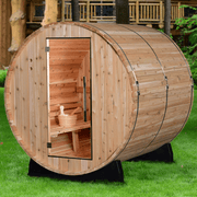 The Almost Heaven Pinnacle 4 Person Barrel Sauna by Almost Heaven features a glass door showing benches and a wooden bucket inside. Set on green grass, it’s surrounded by trees and a wooden structure in the background.