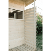 A wooden outdoor shower enclosure with horizontal slats stands by a wood-paneled wall and window on a wooden floor, surrounded by grass and bushes, evoking the inviting feel of the Almost Heaven Timberline 6 Person Cabin Sauna.