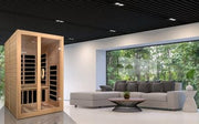 A modern living room with a large gray sectional and coffee table features the Golden Designs Dynamic Santiago 2-person Low EMF FAR Infrared Sauna, crafted from Canadian Hemlock. Floor-to-ceiling windows reveal lush greenery; the black ceiling has recessed lighting.