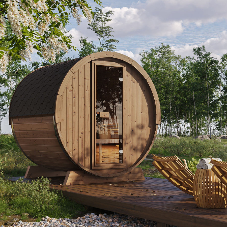 A SaunaLife Model E6W 3 Person Sauna Barrel-Window from SaunaLife rests on a wooden deck amid green trees and grass, with lounge chairs nearby under a partly cloudy sky.