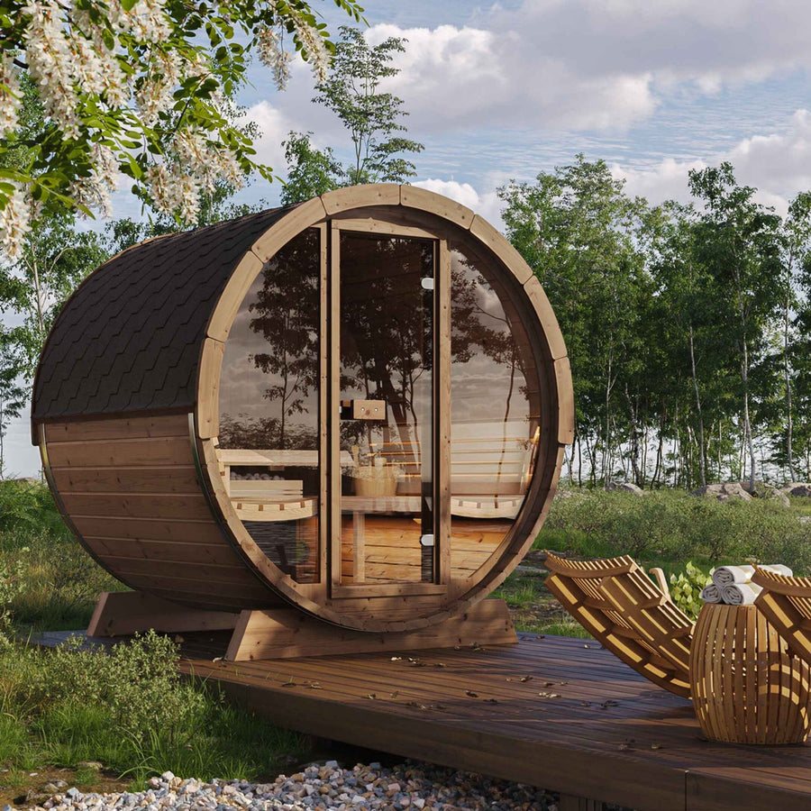 The SaunaLife Model E8 6 Person Barrel Sauna with glass doors is set on a wooden deck among trees, accompanied by two ergonomic lounge chairs and a small table, all beneath a cloudy sky.