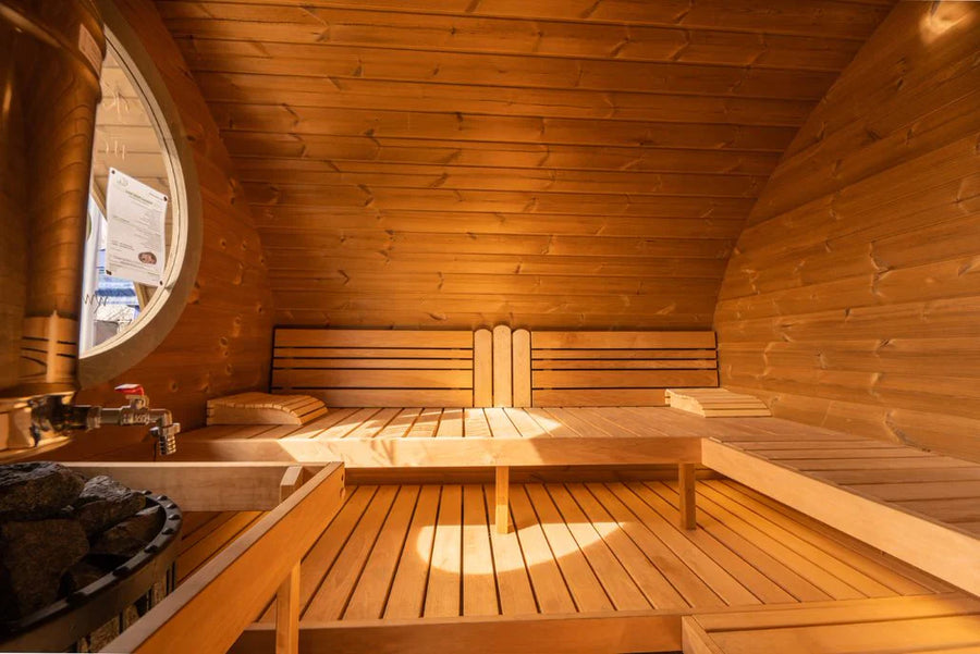 The SaunaLife Model G11 Outdoor 8 Person Sauna features cozy wood benches along curved walls, a sunlit side window, a corner heater with stones, and a comfy pillow—perfect for relaxing in your backyard.