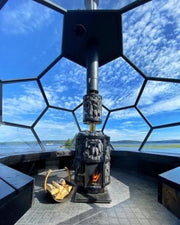 A modern geometric glass-walled room features the Harvia Legend 240 GreenFlame Sauna Stove by Harvia, a log holder with firewood, and panoramic views of blue sky and water outside.