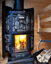 The Harvia Legend 240 GreenFlame Sauna Stove by Harvia burns wood in a wooden-paneled sauna room. Nearby, there’s a stack of firewood and part of a wooden bench is visible on the right.