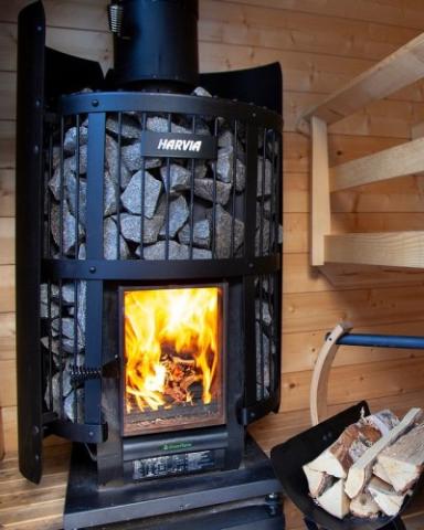 The Harvia Legend 240 GreenFlame Sauna Stove by Harvia burns wood in a wooden-paneled sauna room. Nearby, there’s a stack of firewood and part of a wooden bench is visible on the right.