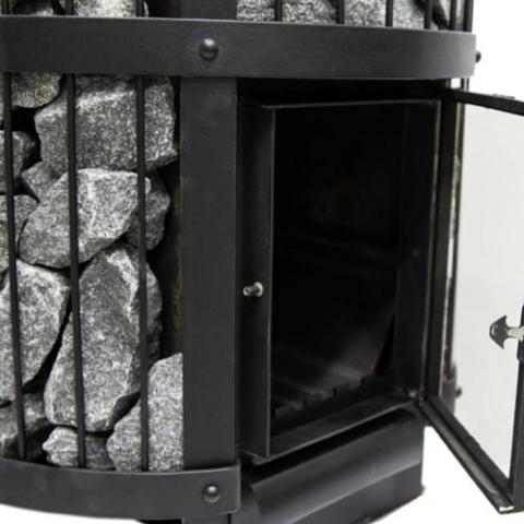 A close-up of a black Harvia Legend 240 GreenFlame Sauna Stove by Harvia shows its side door open, revealing the firebox. Gray stones are stacked vertically behind metal bars along the stove’s edge.