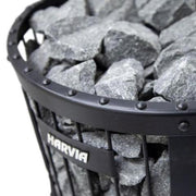 A close-up of a metal basket filled with large grey sauna stones. The black-framed basket, attached to the Harvia Legend 240 GreenFlame Sauna Stove, features the Harvia brand name on a metal plate on the side.
