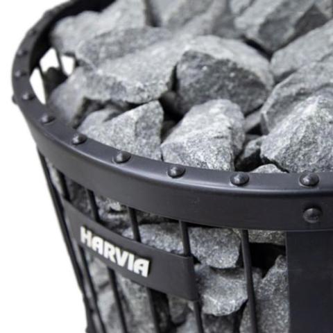 A close-up of a metal basket filled with large grey sauna stones. The black-framed basket, attached to the Harvia Legend 240 GreenFlame Sauna Stove, features the Harvia brand name on a metal plate on the side.