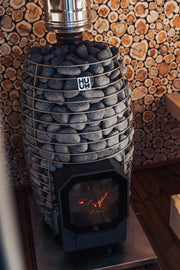 The HUUM Hive Flow LS Wood Burning Sauna Stove by HUUM features a metal frame with smooth gray stones, a glass door revealing the fire, and authentic style enhanced by a backdrop of stacked wood log slices.