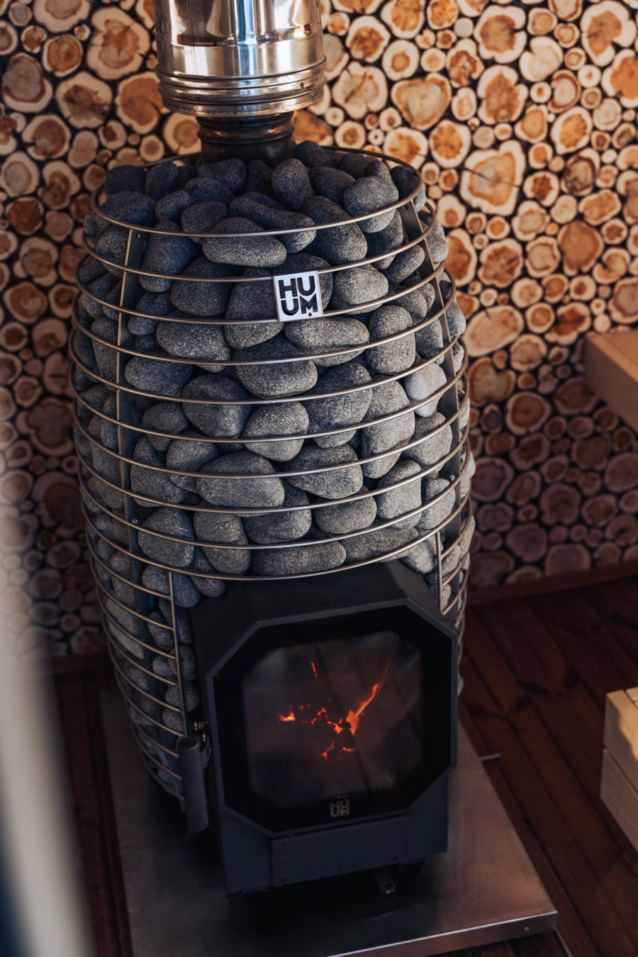 The HUUM Hive Flow LS Wood Burning Sauna Stove by HUUM stands on a metal base, covered with smooth gray stones and glowing with a warm fire, set against a decorative wall with circular log patterns in the background.