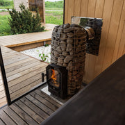 A HUUM Hive Flow LS Wood Burning Sauna Stove glows beside a glass door, with wooden decking and greenery outside. The sauna features wood-paneled walls and floors by HUUM, creating a natural, relaxing atmosphere.