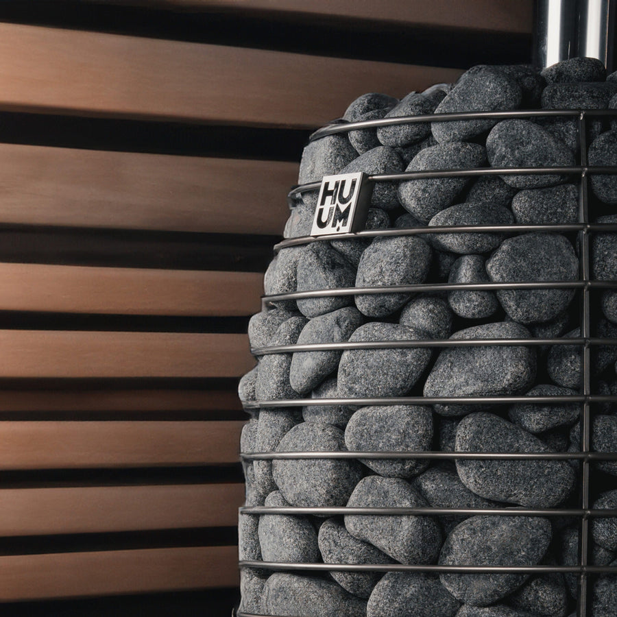 A close-up of the HUUM Hive Flow LS Wood Burning Sauna Stove's metal basket, filled with smooth gray stones and featuring a HUUM tag, set against a backdrop of horizontal wooden slats.