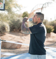 A tattooed man drinks from the MitoHYDRO™ Ultra Premium Hydrogen Water Bottle by Mito Red Light on an outdoor sports court, standing near a net with trees and sunlight in the background.