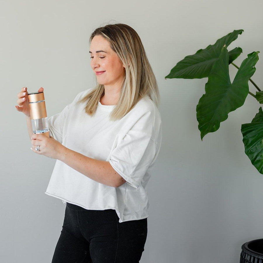 A blonde woman in a white shirt and black pants stands indoors holding the Mito Red Light MitoHYDRO™ Ultra Premium Hydrogen Water Bottle with a gold lid. Behind her, a large green plant sits against a gray wall.