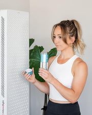 A woman in a white crop top holds a MitoHYDRO™ Ultra Premium Hydrogen Water Bottle by Mito Red Light and a cup, standing next to a tall white red light therapy device with a grid of lights. A leafy green plant is in the background.