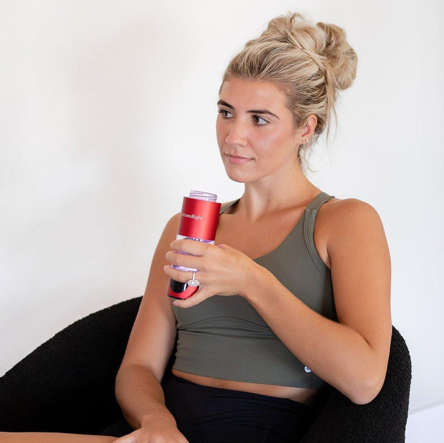 A woman with blonde hair in a messy bun, wearing a green sports bra and black shorts, sits on a black chair holding the MitoHYDRO™ Ultra Premium Hydrogen Water Bottle by Mito Red Light, looking to the side with a neutral expression.