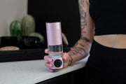 A person with colorful tattoos and pink nail polish holds a MitoHYDRO™ Ultra Premium Hydrogen Water Bottle by Mito Red Light on a white kitchen counter, with cacti and black containers visible in the background.