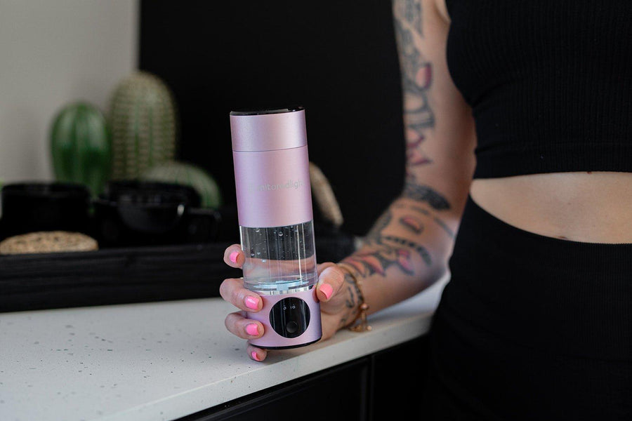 A person with colorful tattoos and pink nail polish holds a MitoHYDRO™ Ultra Premium Hydrogen Water Bottle by Mito Red Light on a white kitchen counter, with cacti and black containers visible in the background.