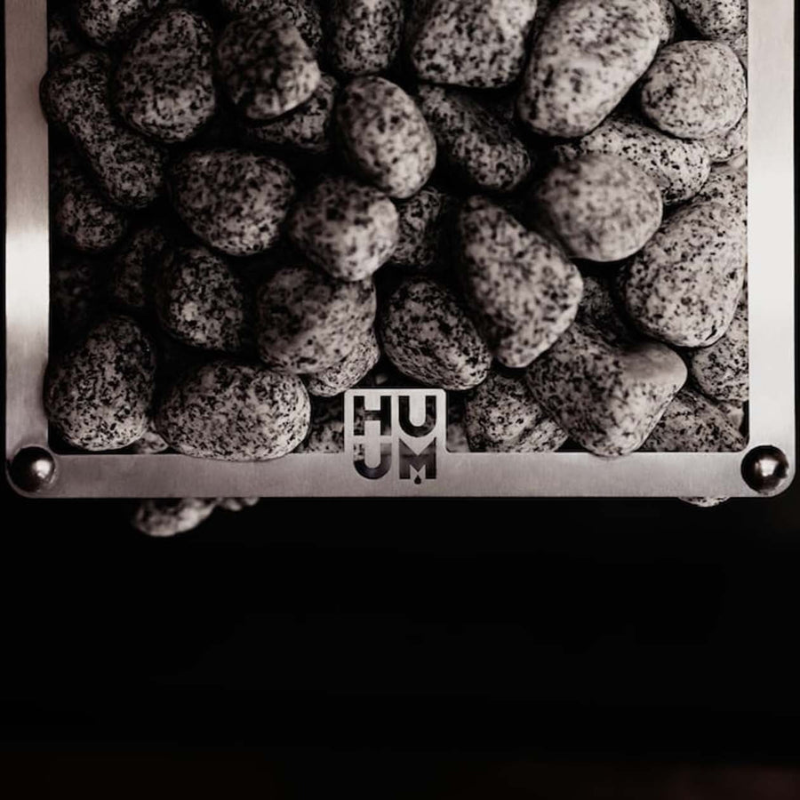A close-up of gray stones stacked inside a metal frame with "HUUM" cut out in the center, highlighting the modern design of the HUUM CLIFF Series 11kW Sauna Heater Package by HUUM.