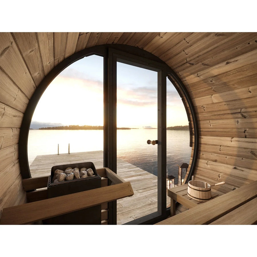 Inside the SaunaLife Model EE6G 4 Person Barrel Sauna, a glass door offers views of a dock over a tranquil lake at sunset, while a sauna heater with stones and a wooden bucket add to this cozy SaunaLife retreat.
