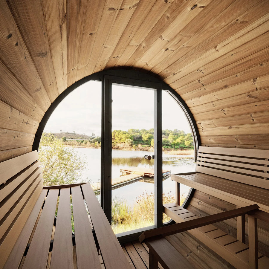 Interior of the SaunaLife Model EE8G 6 Person Barrel Sauna with benches, a large round window, and a glass door overlooking a lake, dock, and lush trees—a perfect backyard retreat from SaunaLife.