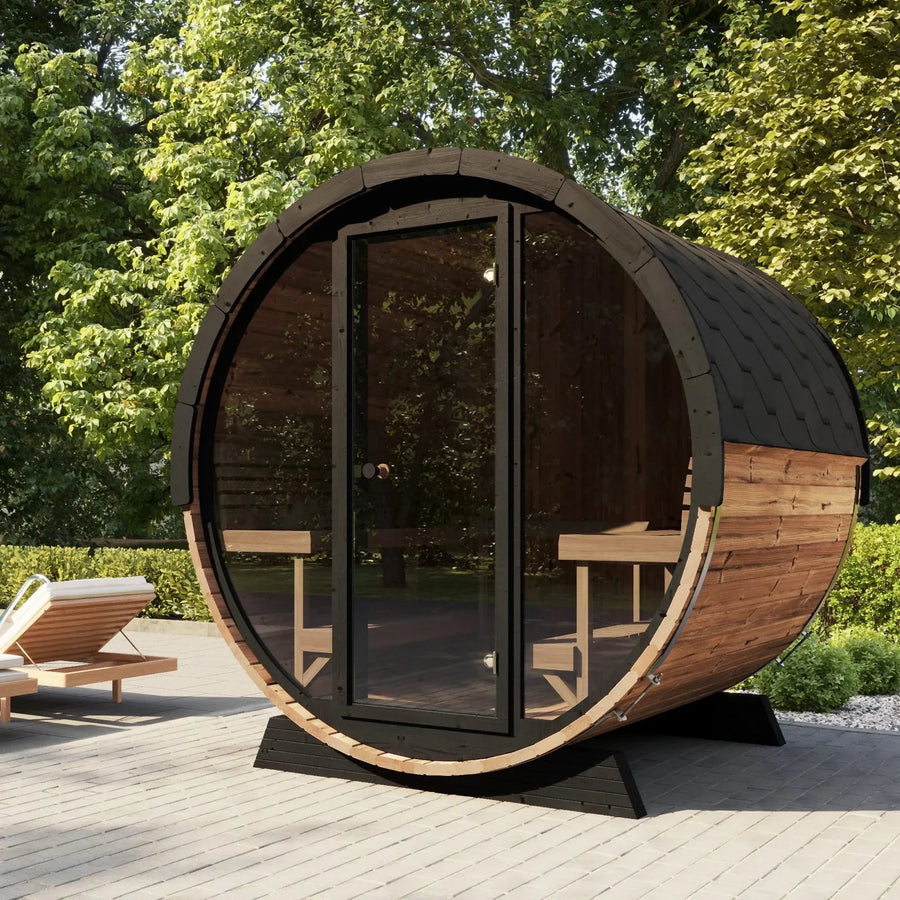 The SaunaLife Model EE8G 6 Person Barrel Sauna by SaunaLife, featuring a glass door, sits on a patio among lush greenery. A nearby lounge chair with a white cushion offers a perfect spot to relax in the sunlit space.
